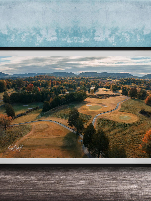 The image is a framed aerial view of a golf course surrounded by trees and rolling hills, with a blue sky and clouds overhead.