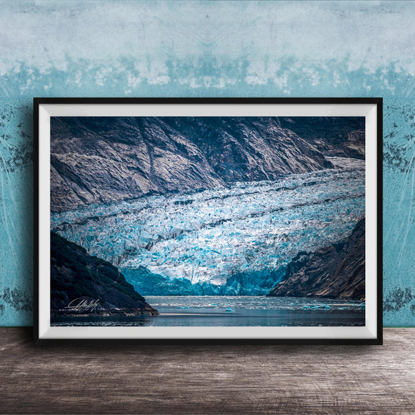 A framed photograph of a large, majestic glacier with a blue-green hue, set against a backdrop of rugged mountains and a serene body of water.