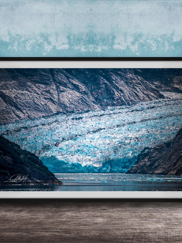 A framed photograph of a large, majestic glacier with a blue-green hue, set against a backdrop of rugged mountains and a serene body of water.