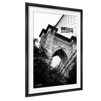 A black and white photograph of a brick bridge with an American flag flying above it, framed in a black frame.