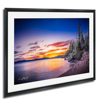 A framed photograph depicting a serene landscape with a calm lake reflecting the vibrant colors of a sunset sky, surrounded by trees and mountains.