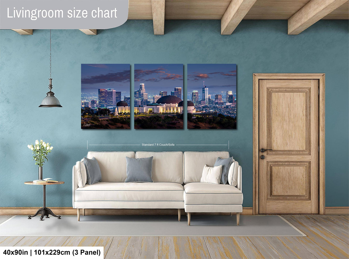 The image is a living room with a large wall art depicting a city skyline at night. The wall art is divided into three panels, each showing a different part of the cityscape. The room has a white sofa, a wooden door, and a small table with a vase of flowers. The wall art is approximately 40 inches wide and 101.29 cm tall, with a 3-panel layout.