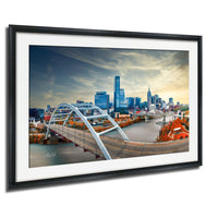 The image is a framed photograph of a city skyline, featuring a large bridge spanning a river with a colorful autumnal backdrop.