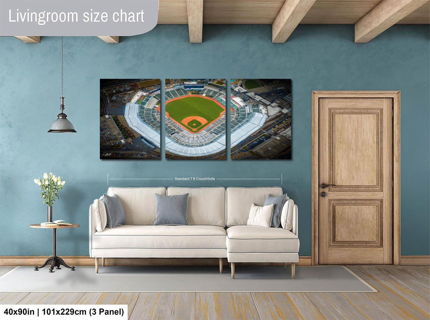 The image is a living room with a large wall-mounted painting depicting an aerial view of a baseball stadium. The painting is divided into three panels, each showing different parts of the stadium. The room has a white sofa, a wooden door, and a small table with a vase of flowers. The painting is approximately 40 inches wide and 101.29 cm tall, with a 3-panel layout.