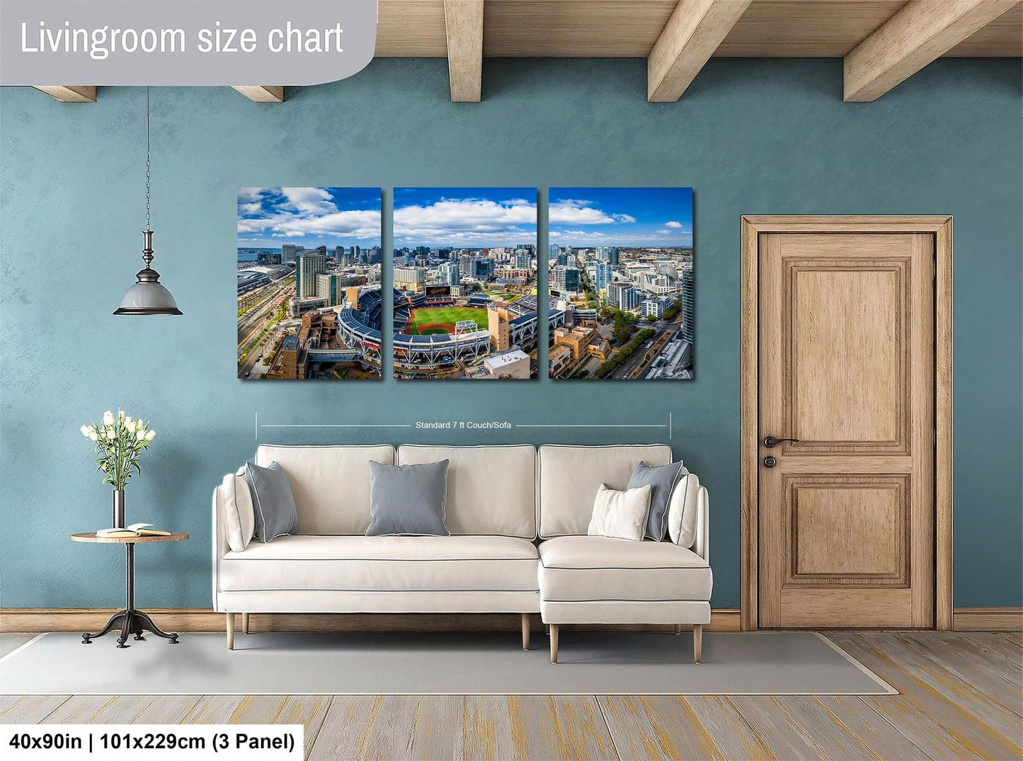 The image is a living room with a large wall-mounted painting depicting a cityscape. The painting is divided into three panels, each showing a different part of the city. The room has a white sofa, a wooden door, and a small table with a vase of flowers. The painting is approximately 40 inches wide and 101.29 cm tall, with a 3-panel arrangement.