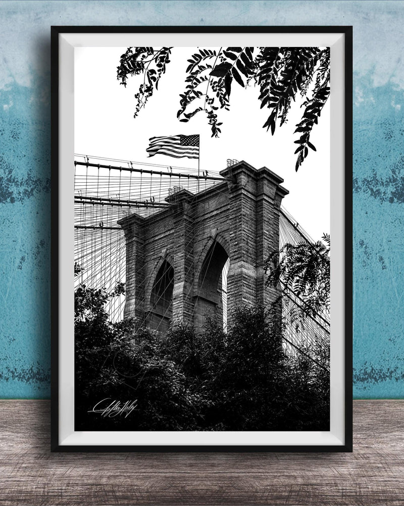 A black and white photograph of the Brooklyn Bridge, with the American flag flying atop the structure.