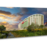 A large stadium with a curved roof and a bridge in the foreground, set against a colorful sky with clouds.