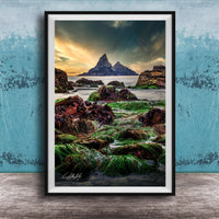 The image is a framed photograph of a rocky shoreline with a large, distant mountain in the background. The photograph is set against a blue wall, and the sky above is cloudy. The image is a collage of multiple images, and the photograph is framed with a black border.