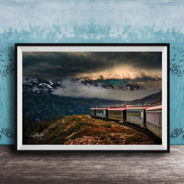 A framed photograph depicting a train traveling through a mountainous landscape with dramatic clouds and a blue sky in the background.