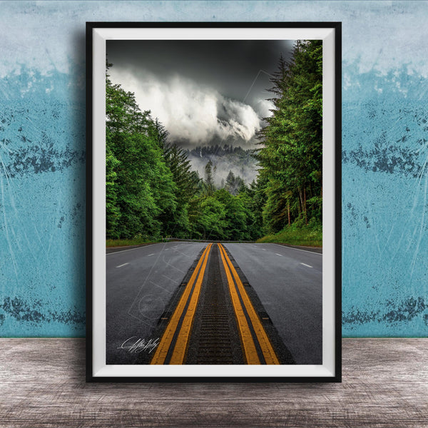 A framed photograph depicting a winding road surrounded by lush green trees and a dramatic cloudy sky in the background.