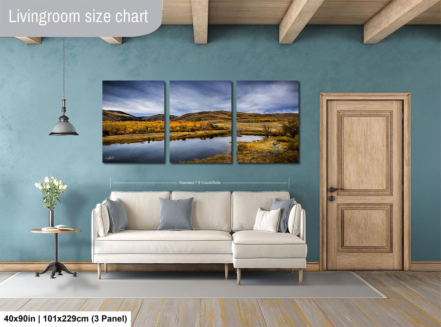 The image is a living room with a large wall art depicting a scenic landscape with a lake and mountains. The living room has a white sofa and a wooden door, and there is a potted plant and a vase on a side table. The wall art is approximately 40 inches wide and 101/129 centimeters tall, and it is a three-panel collage.