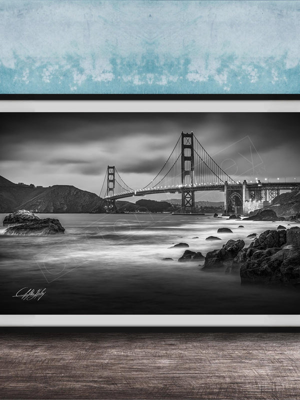 A black and white photograph of the Golden Gate Bridge in San Francisco, California, with the bridge's towers and suspension cables prominently featured against a cloudy sky.