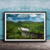 A framed photograph depicting a scenic train traveling over a stone bridge in a lush, green landscape with rolling hills and a blue sky with clouds.