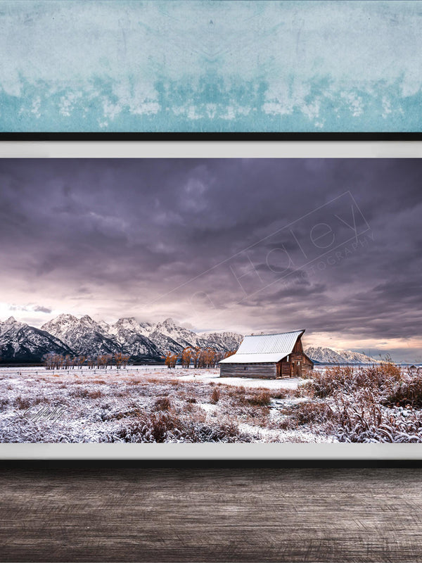 A framed photograph depicting a snowy mountain landscape with a wooden barn in the foreground, set against a cloudy sky.