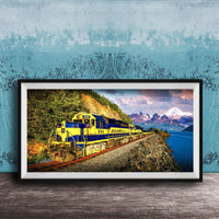 A framed photograph of a yellow and blue train traveling along a track through a mountainous landscape with a body of water in the background.