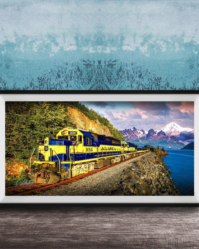 A framed photograph of a yellow and blue train traveling along a track through a mountainous landscape with a body of water in the background.