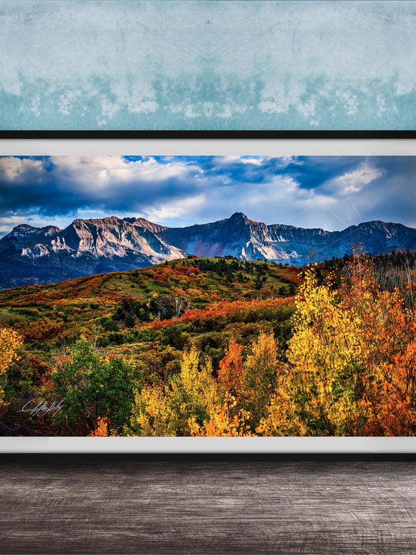 The image is a framed photograph of a mountainous landscape with autumn-colored trees in the foreground and a blue sky with clouds in the background.