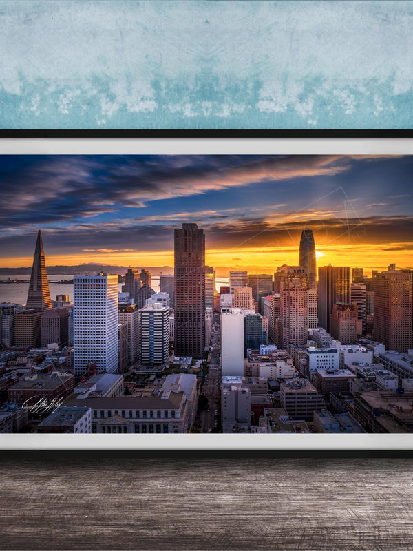The image is a framed photograph of a city skyline at sunset, with the sun setting behind the buildings and casting a warm, golden glow over the urban landscape.