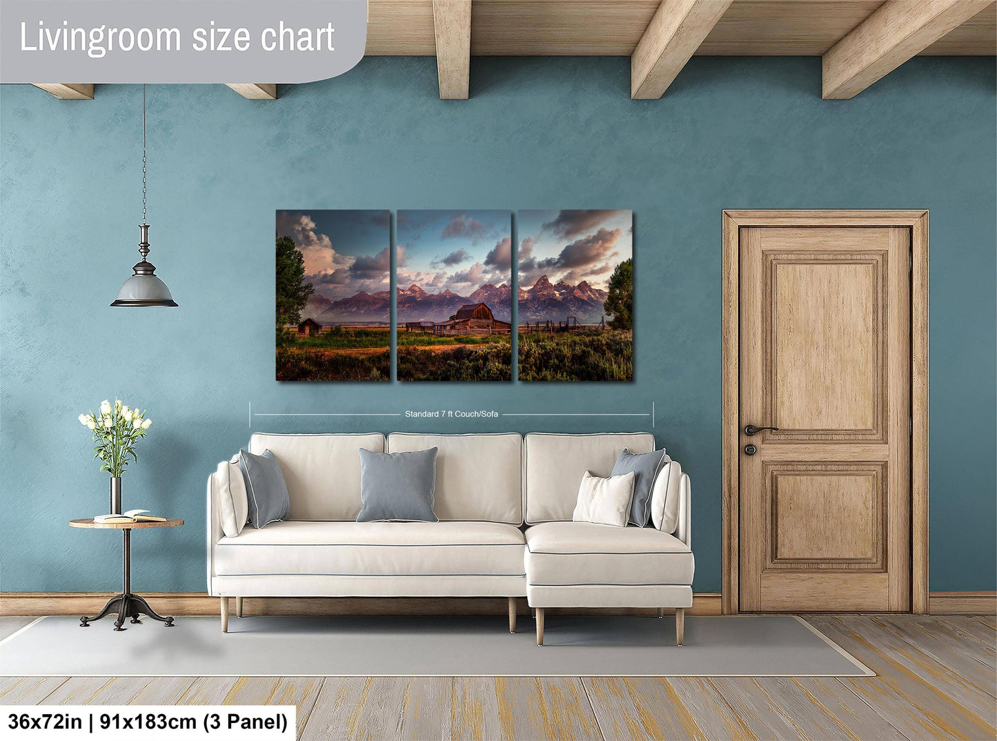 The image is a living room with a large, three-panel landscape painting hanging on the wall. The painting depicts a scenic mountain landscape with a sunset. The room has a white sofa and a wooden door, and there is a small table with a vase of flowers nearby. The painting is approximately 36 inches wide and 15 inches tall, and it is mounted on a wooden beam.