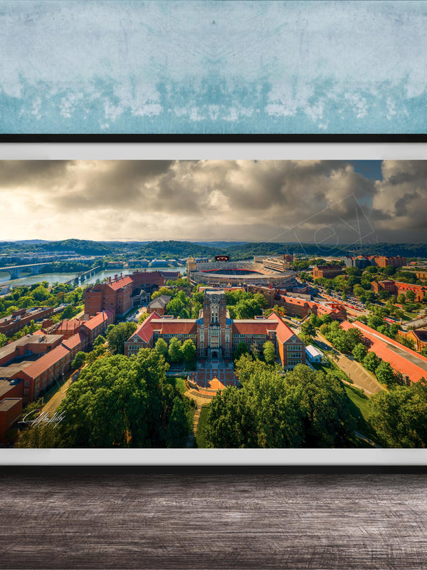 The image is a framed aerial view of a city with a mix of buildings, trees, and a river. The sky is cloudy, creating a dramatic backdrop for the cityscape.