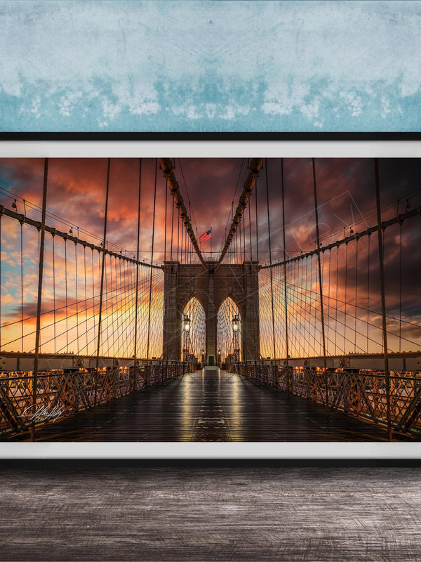 A framed photograph of the Brooklyn Bridge at sunset, with the bridge's intricate metal structure and suspension cables silhouetted against a vibrant sky filled with clouds and warm hues.