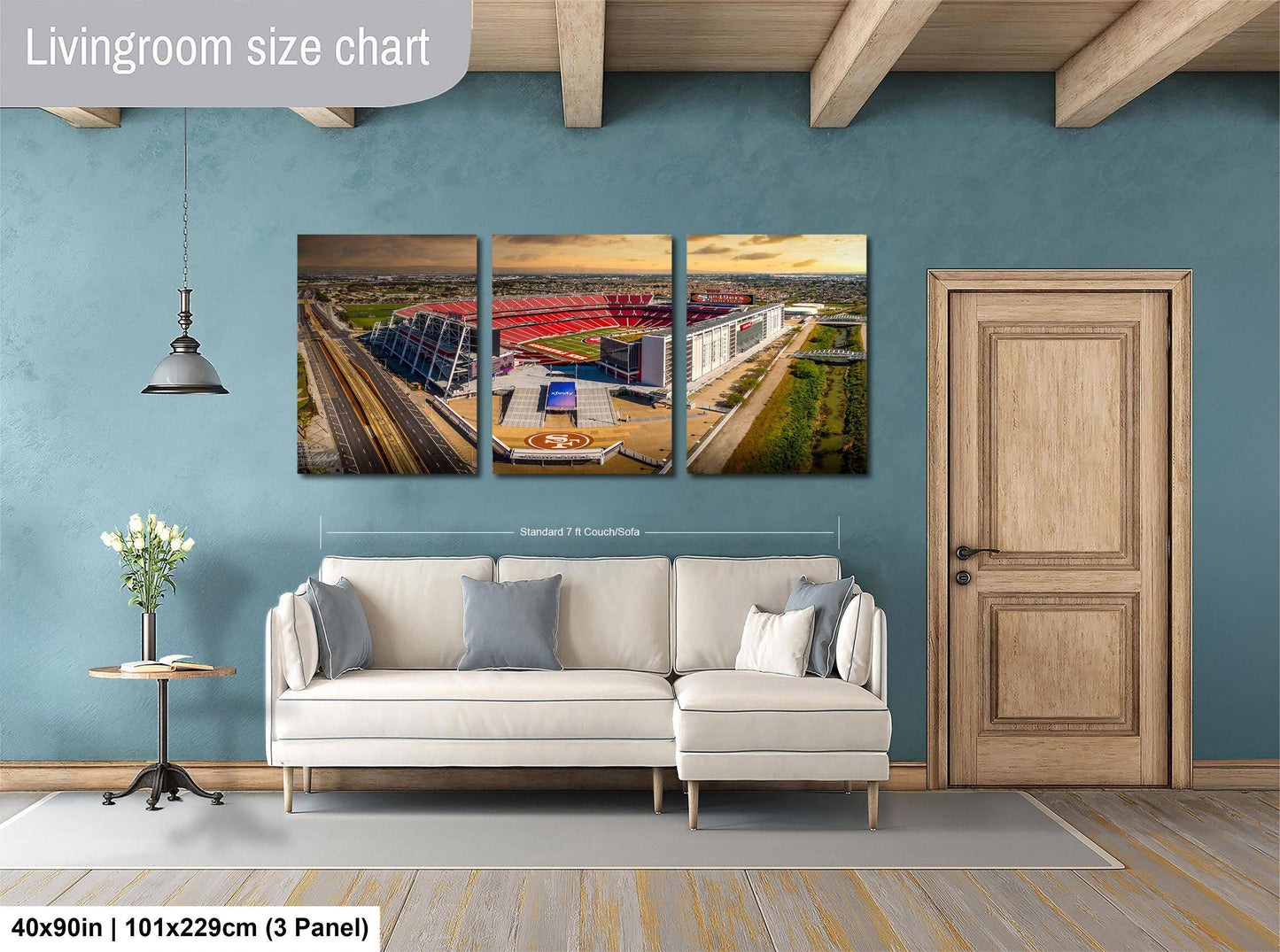 The image is a living room with a large wall art piece depicting a stadium or sports arena. The living room has a white sofa, a wooden door, and a wooden floor. The wall art is approximately 40 inches wide and 101.29 cm tall, with a 3-panel layout.