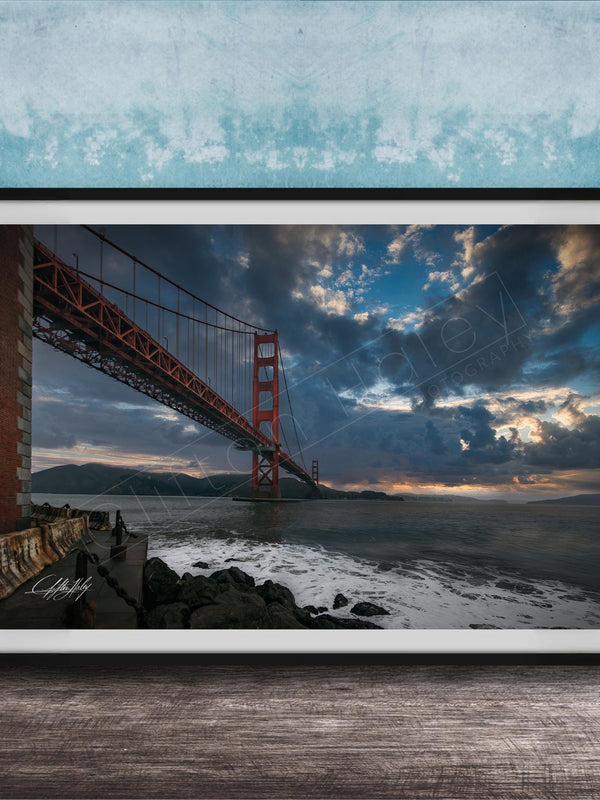A framed photograph of the Golden Gate Bridge in San Francisco, California, with dramatic clouds and a sunset sky in the background.