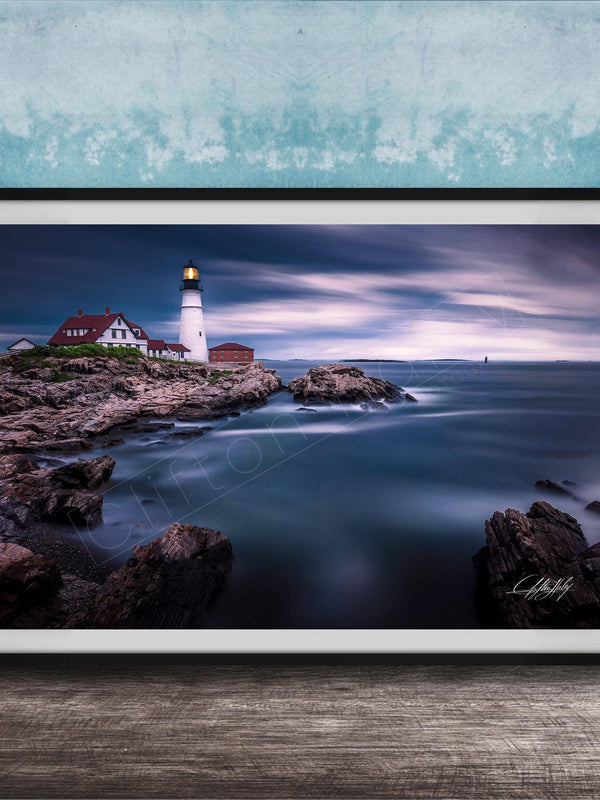 A framed photograph depicting a lighthouse on a rocky coastline, with the ocean and a cloudy sky in the background.