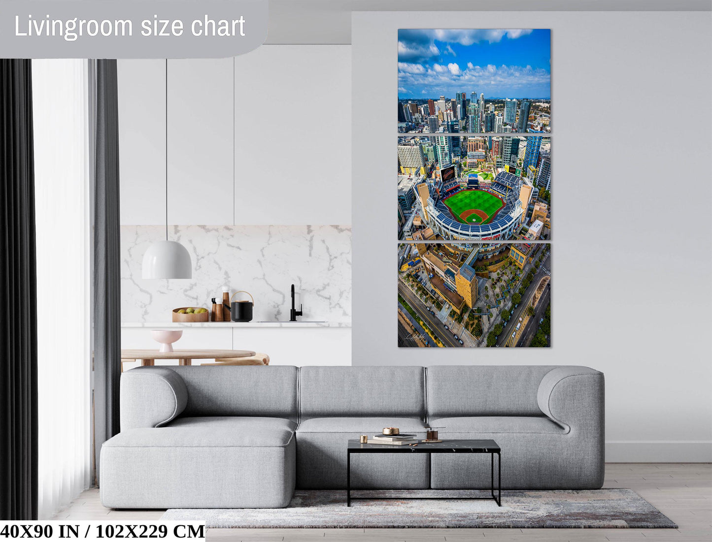 The image is a living room with a gray sofa and a coffee table. Above the sofa, there is a large wall art depicting an aerial view of a city skyline, including a baseball stadium.
