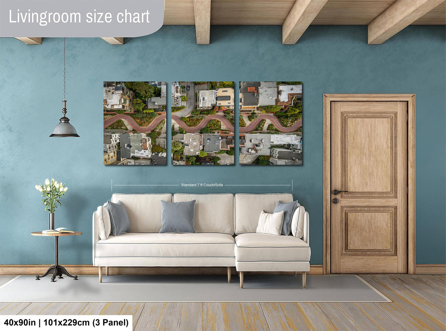 The image is a living room with a large wall art depicting an aerial view of a cityscape. The living room has a white sofa and a wooden door, and there is a potted plant and a lamp on a side table. The wall art is approximately 40 inches wide and 101.29 cm in height, and it is divided into three panels.