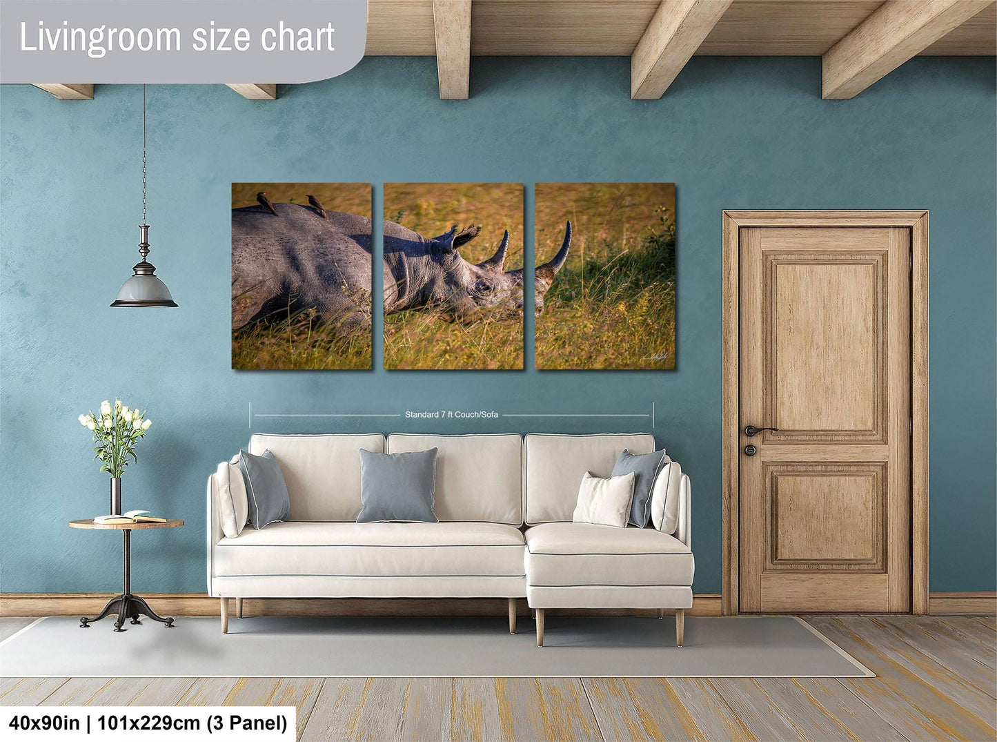 The image is a living room with a large white sofa, a wooden door, and a hanging light fixture. Above the sofa, there is a wall-mounted painting depicting a rhinoceros in a grassy field. The painting is divided into three panels, with the largest panel in the center and the smaller ones on either side. The wall behind the sofa has a blue color, and the floor is covered with a gray rug.