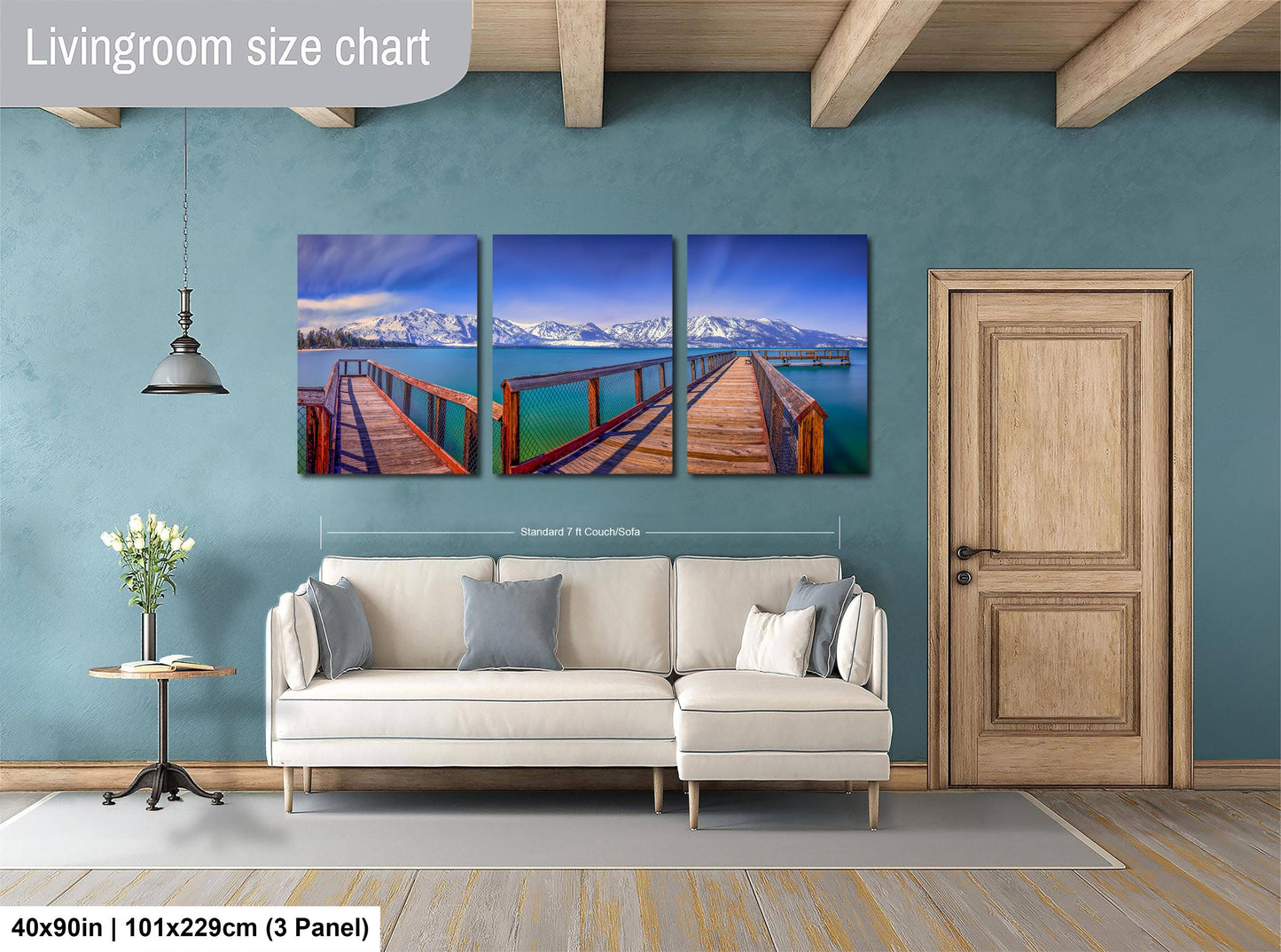 The image is a living room with a large white sofa, a wooden door, and a wooden beam ceiling. Above the sofa, there is a wall-mounted living room size chart depicting a scenic view of a wooden bridge over a body of water with mountains in the background.
