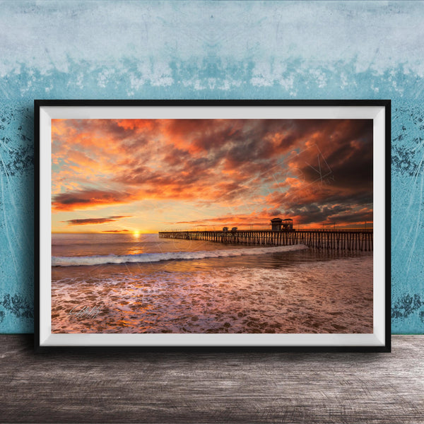 A framed photograph depicting a serene beach scene at sunset, with a wooden pier extending into the water and the sky filled with vibrant orange and pink hues.