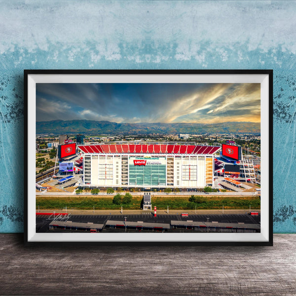 A framed photograph of a large sports stadium with a red roof, surrounded by mountains and a cloudy sky.