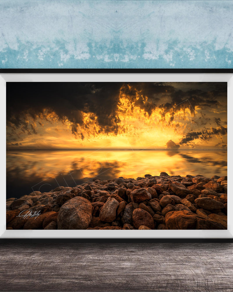 The image is a framed photograph depicting a rocky shoreline at sunset, with the sky filled with vibrant orange and yellow hues reflecting on the calm water surface.