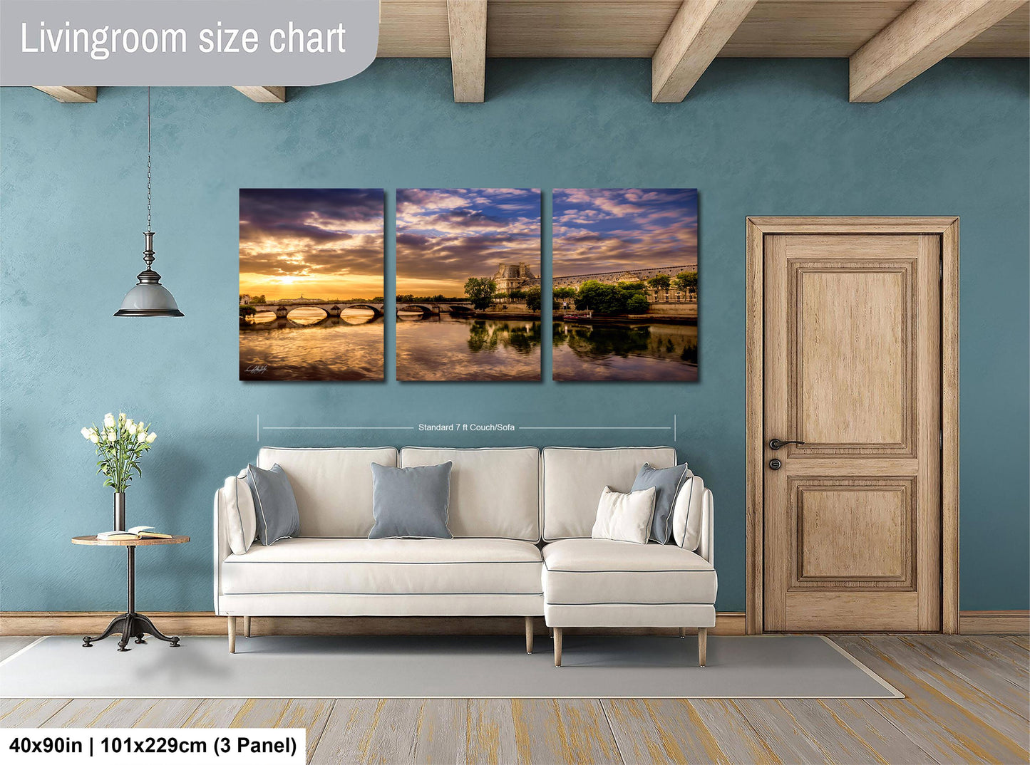 The image is a living room with a large wall art panel depicting a scenic landscape with a river, bridge, and sunset. The living room has a white sofa, a wooden door, and a small table with a vase of flowers. The wall art panel is approximately 40 inches wide and 101.29 cm tall, and it is a 3-panel design.