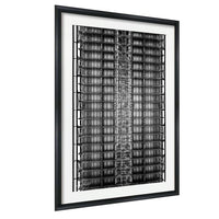 The image is a black and white photograph of a tall, intricate metal structure with numerous levels and rows of bars or beams. The photograph is framed and displayed on a wall.