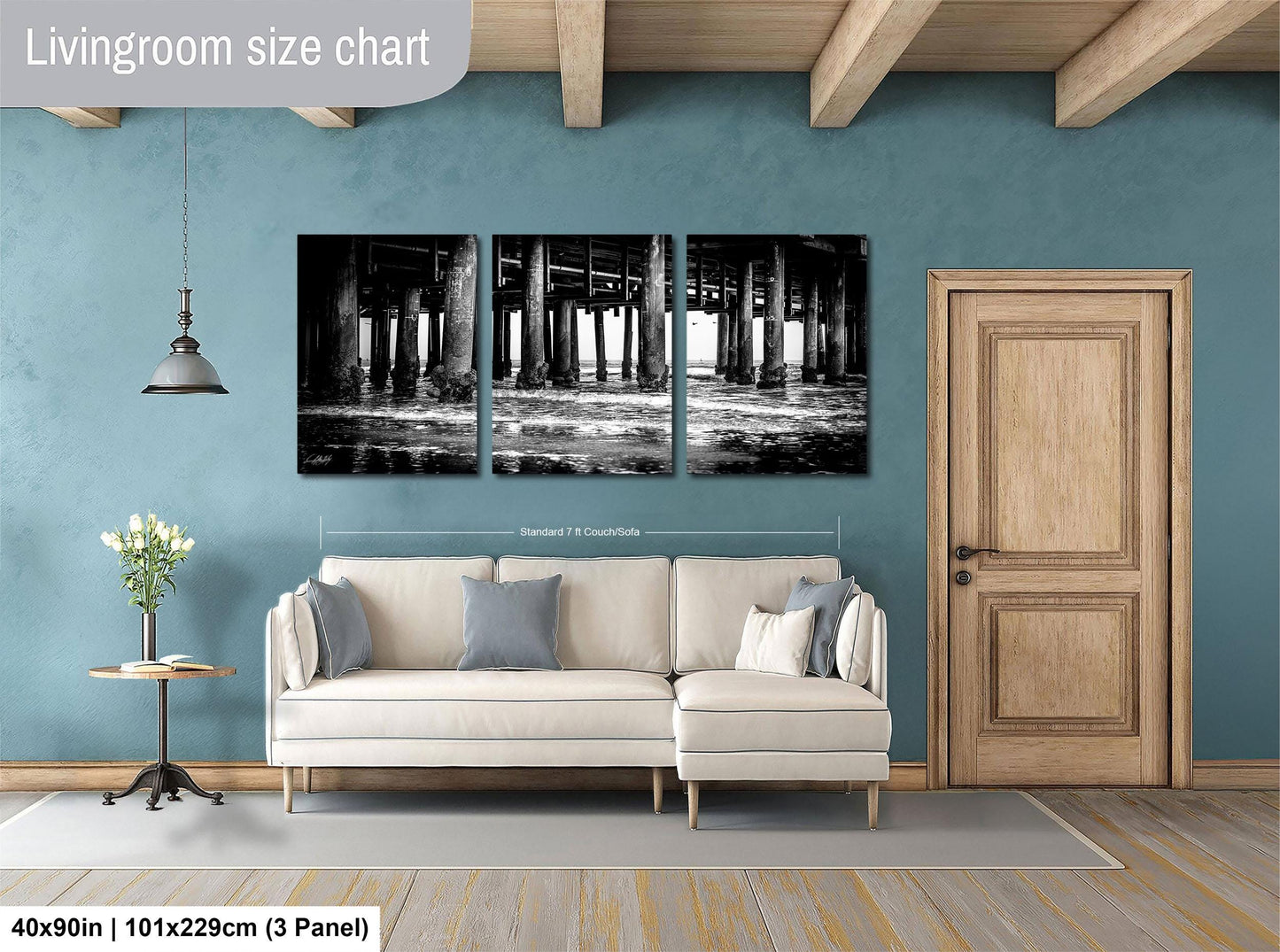 The image is a living room with a large, three-panel black and white photograph of a bridge hanging on the wall. The photograph is framed in a standard size of 71 x 129 cm (3 x 3 panel). The room also includes a white sofa, a wooden door, and a small table with a vase of flowers.