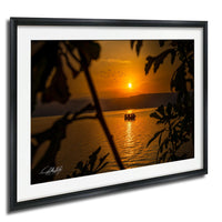 A framed photograph depicting a serene sunset over a body of water, with silhouetted trees framing the scene.