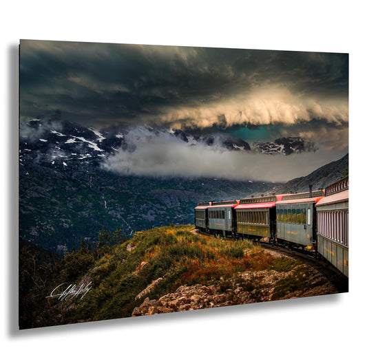 A train travels through a mountainous landscape, with snow-capped peaks and a dramatic cloudy sky in the background.
