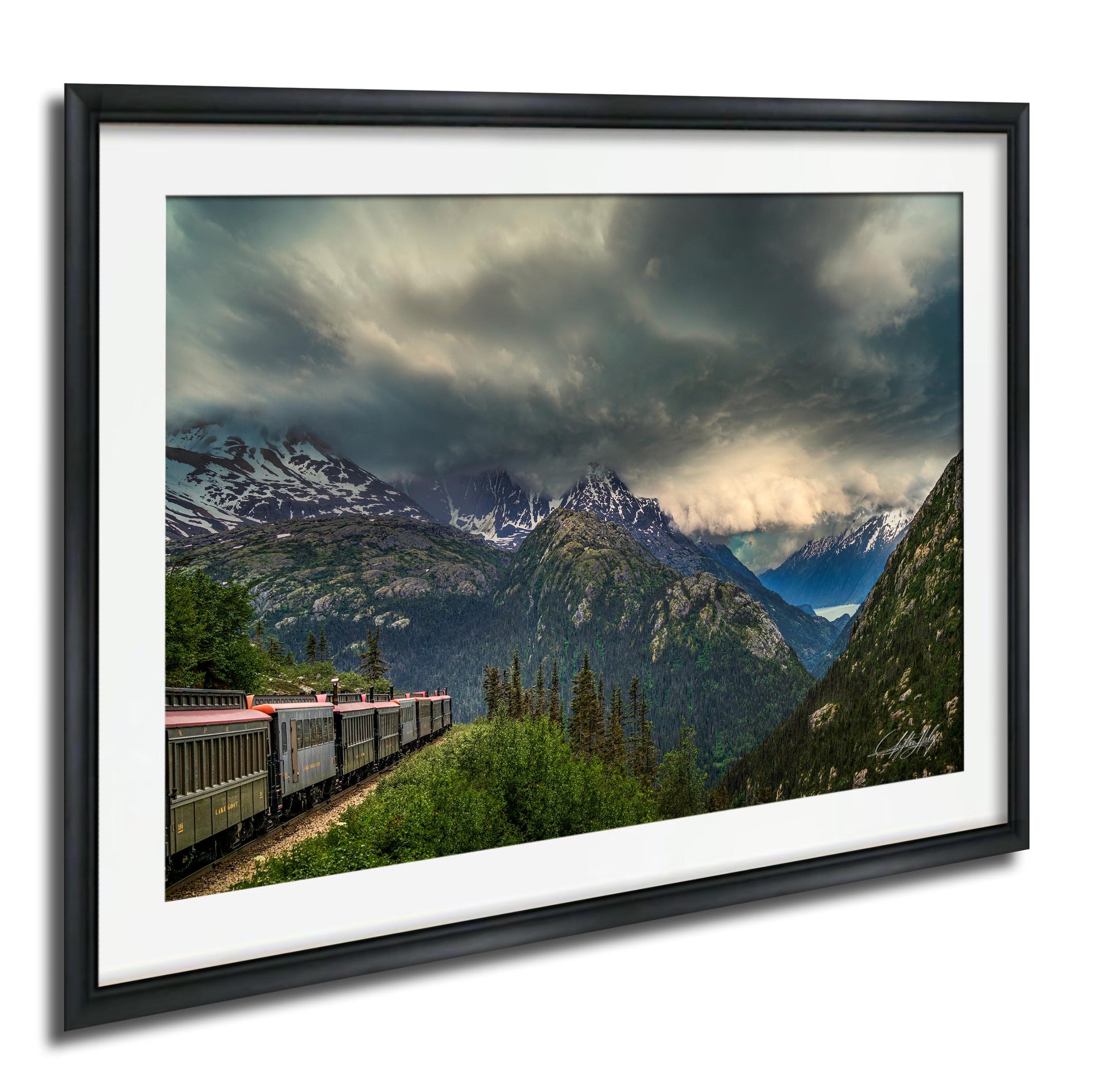 A framed photograph depicting a train traveling through a mountainous landscape with snow-capped peaks and a dramatic cloudy sky.