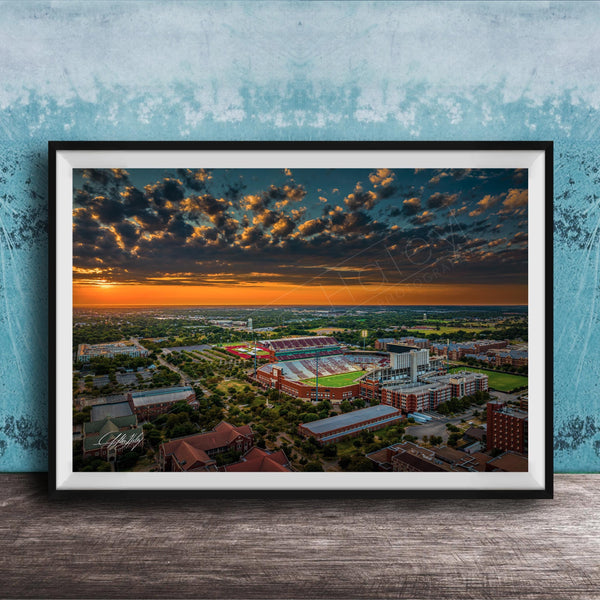The image is a framed aerial view of a cityscape at sunset, with the sky filled with clouds and the sun setting over the horizon.
