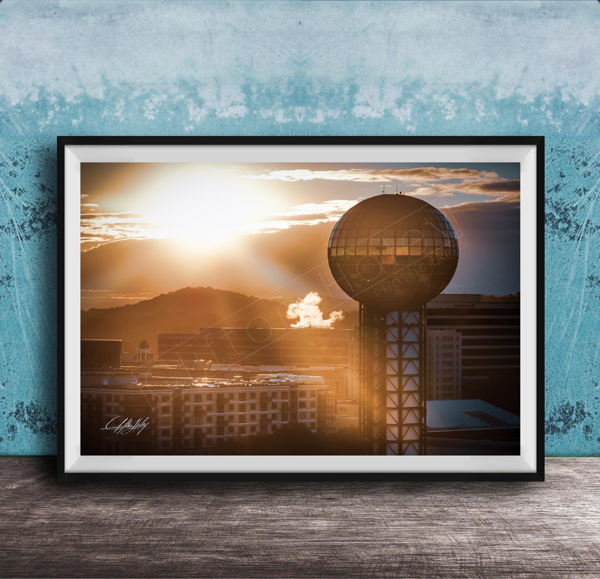 A framed photograph of a city skyline at sunset, with a large tower in the foreground and the sun setting behind the buildings.