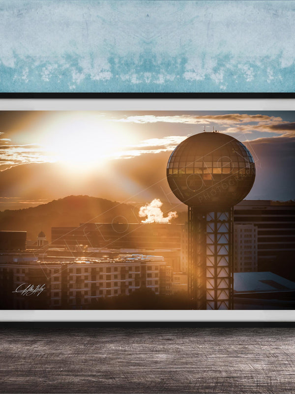 A framed photograph of a city skyline at sunset, with a large tower in the foreground and the sun setting behind the buildings.