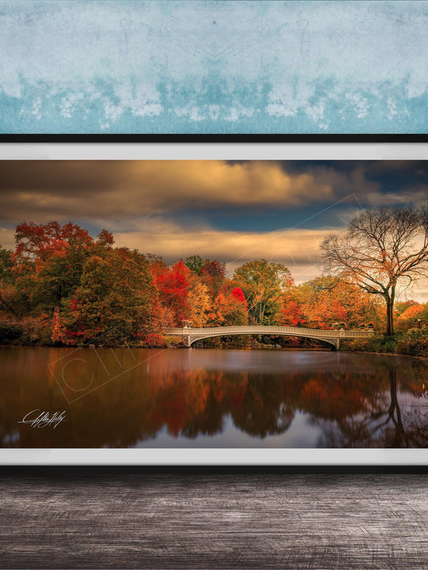 A framed photograph depicting a scenic autumn landscape with a bridge, trees, and a body of water reflecting the vibrant colors of the season.