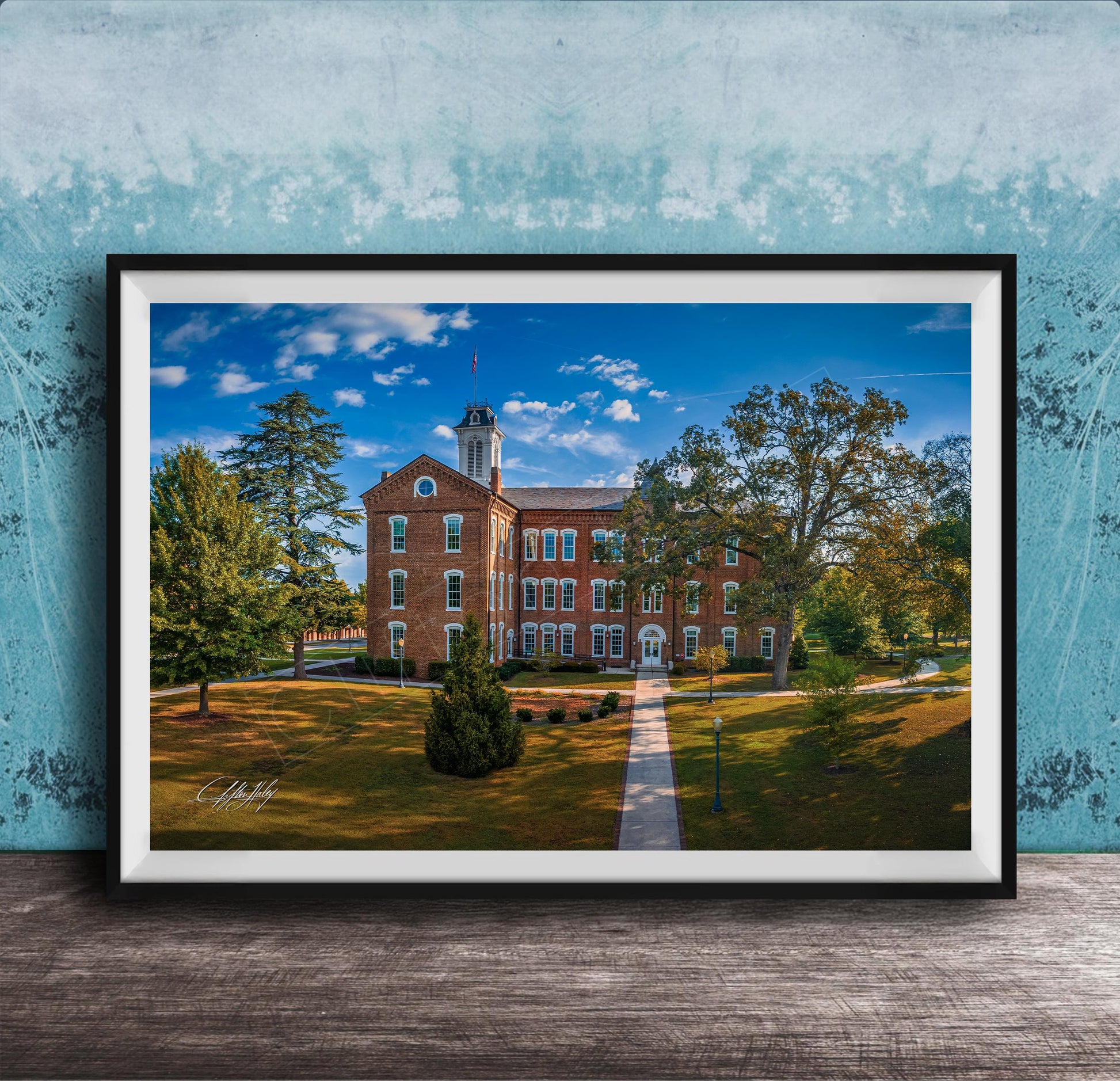 A framed photograph of a brick building with a tower, surrounded by trees and a grassy area, set against a blue sky with clouds.