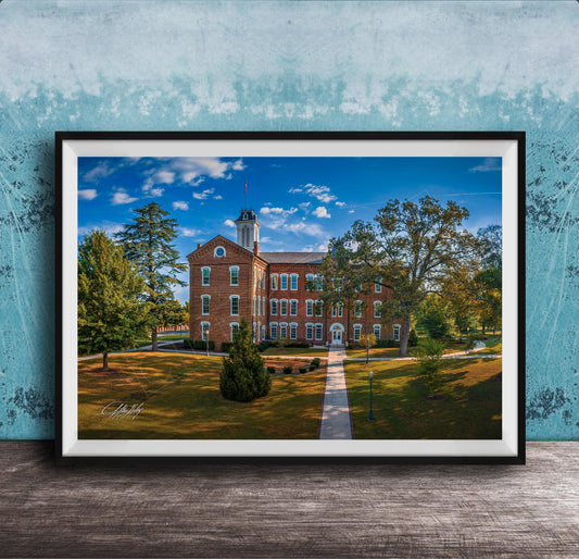 A framed photograph of a brick building with a tower, surrounded by trees and a grassy area, set against a blue sky with clouds.