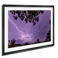 A framed photograph depicting a dramatic lightning storm with bright bolts of light illuminating the sky and silhouetting trees in the foreground.