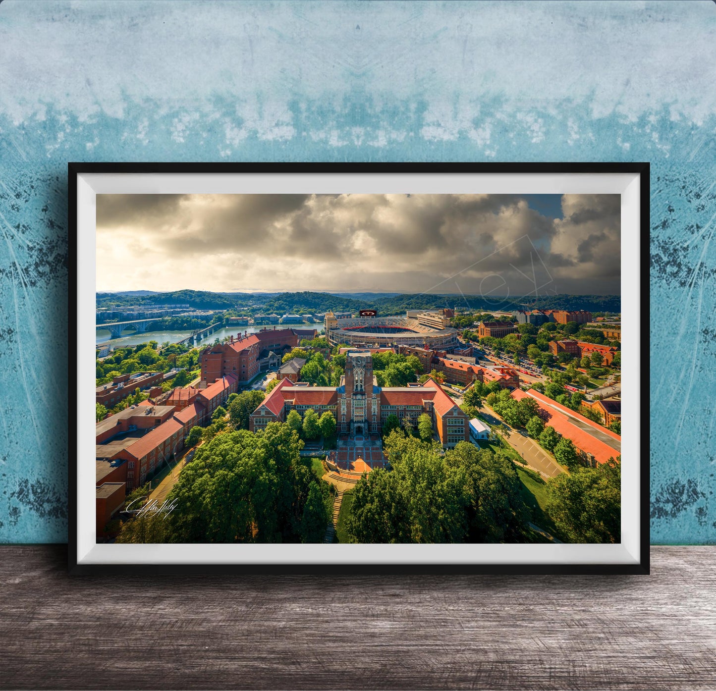 The image is a framed aerial view of a city with a mix of buildings, trees, and a river. The sky is cloudy, creating a dramatic backdrop for the cityscape.
