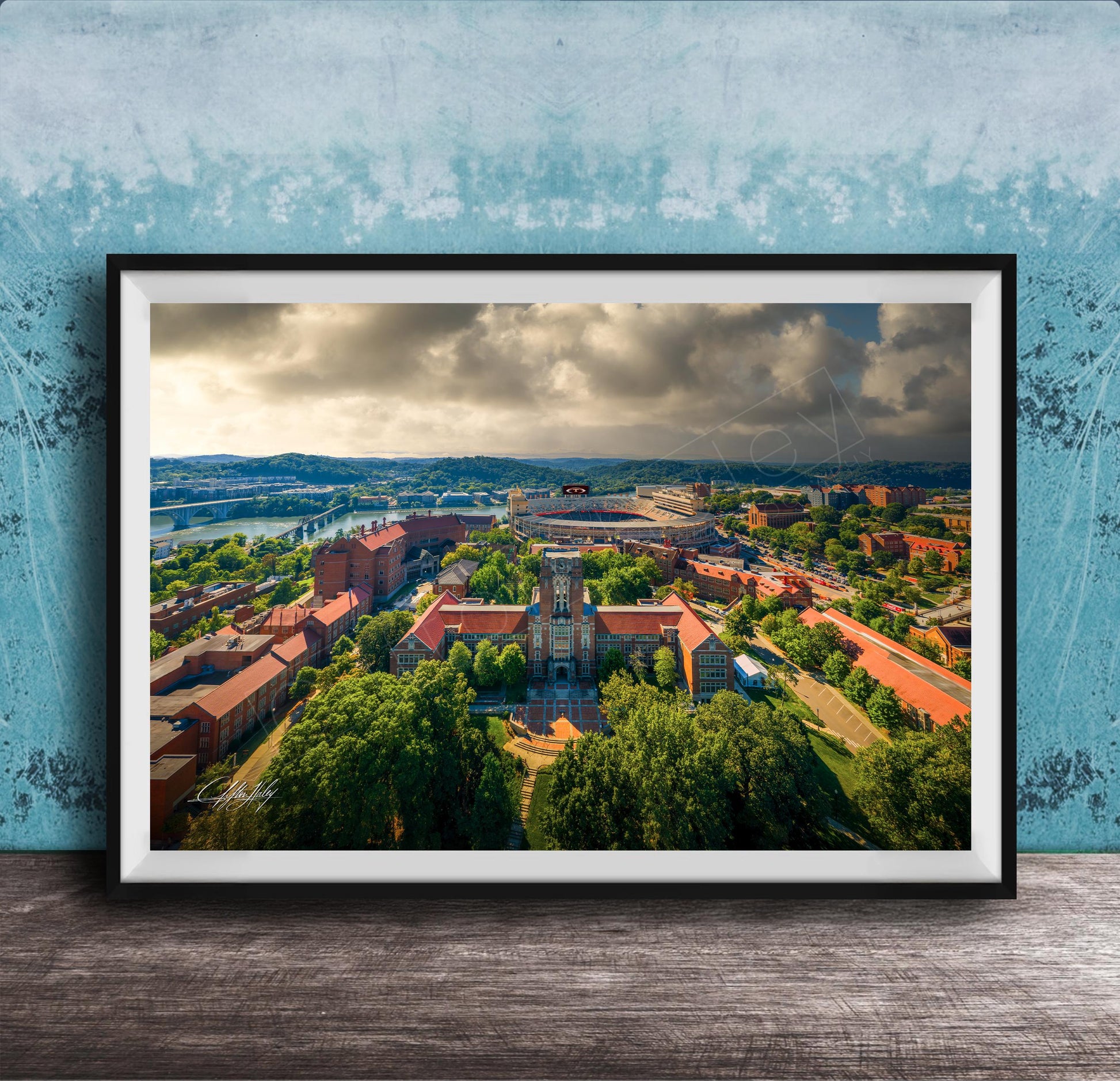 The image is a framed aerial view of a city with a mix of buildings, trees, and a river. The sky is cloudy, creating a dramatic backdrop for the cityscape.